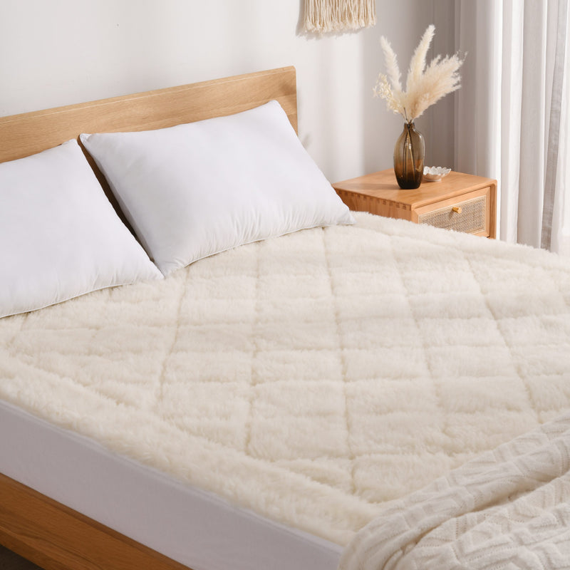 Cozy bed with white pillows and a fluffy cream quilted blanket, next to a wooden nightstand with a vase