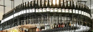 Hanging wine rack with rows of wine bottles on top and upside-down wine glasses below in a bar setting.