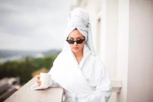 Woman in a white bathrobe and towel turban wearing sunglasses, holding a cup on a balcony