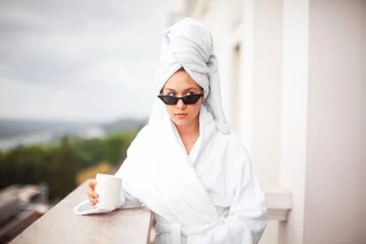 Woman in a white bathrobe and towel turban wearing sunglasses, holding a cup on a balcony