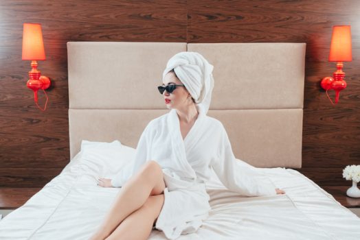 Woman beauty. Female in bathrobe posing on bed. Bare legs. Sunglasses and towel turban on. Luxury hotel room interior.